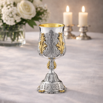 Decorative silver and gold goblet on a table with candles and flowers in the background